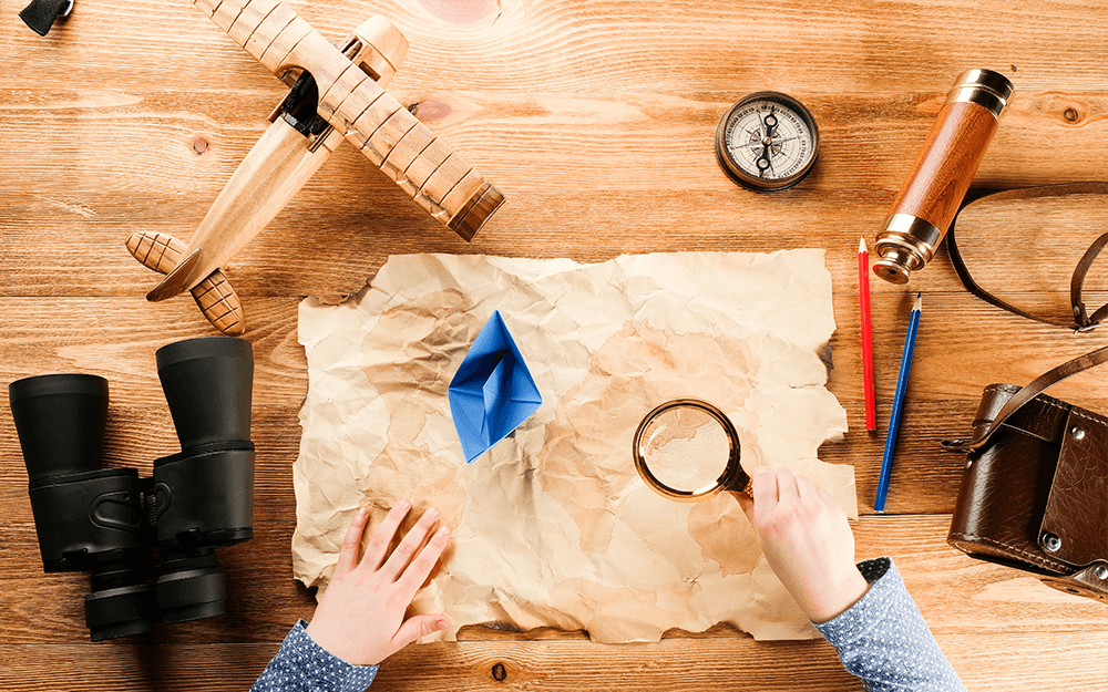 A child holding a magnifying glass, below is a paper with his toys like wooden airplane, telescope, compass, and some pencils.