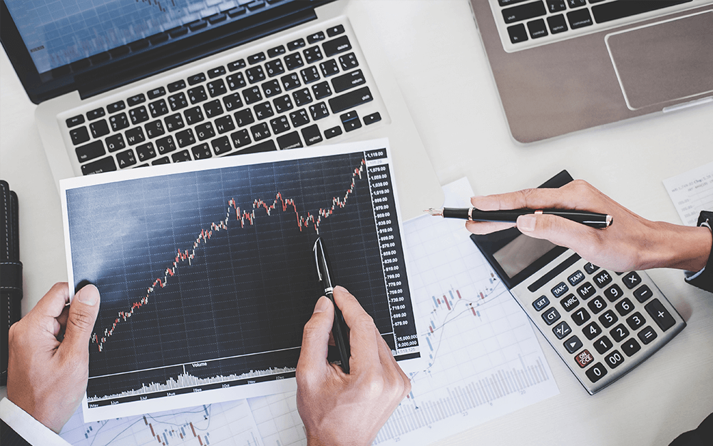 2 men working trading technical analysis holding a paper and pen, and using a laptop and computer.